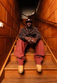 Άνδρας που φοράει καφέ φλις μπουφάν, κόκκινο καρό παντελόνι και κίτρινα μπότες, καθισμένος σε ξύλινη σκάλα σε έναν στενό διάδρομο με ξύλινη επένδυση.