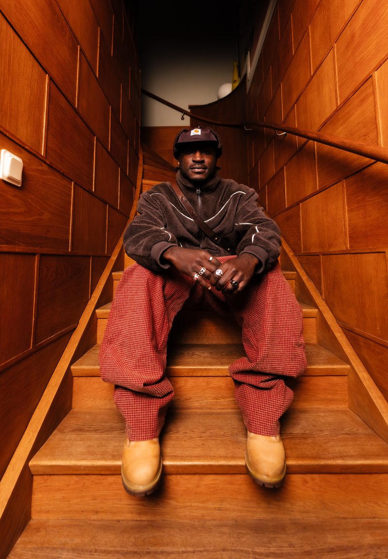 Muž v hnedej fleecovej bunde, červených kockovaných nohaviciach a žltých topánkach sediaci na drevených schodoch v úzkej chodbe obloženej drevom.