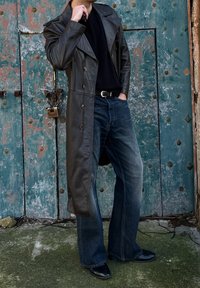 Άτομο που φορά μαύρο δερμάτινο παλτό τύπου τρανς, μαύρο πουκάμισο, μπλε τζιν και μαύρα παπούτσια, στέκεται μπροστά σε μια φθαρμένη μπλε-πράσινη μεταλλική πόρτα.