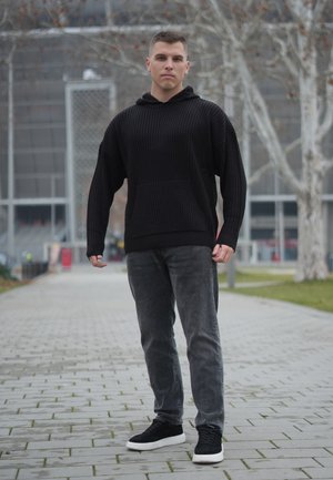 Sudadera negra acanalada con capucha y bolsillo canguro, combinada con vaqueros grises y zapatillas negras, sobre un camino pavimentado con árboles al fondo.