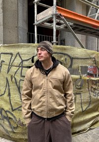 Jaqueta bomber bege com fecho à frente, usada por cima de um hoodie preto. Calças de fato de treino castanho-escuro. Gorro de camuflagem. Fundo urbano com uma lona.