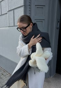 Άσπρο παλτό με υφή και oversized γραμμή, συνδυασμένο με σκοτεινό γκρι κασκόλ. Κρατάει μια αφράτη φωτεινή τσάντα. Γυαλιά με μαύρο πλαίσιο.