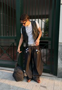 Gilet en cuir noir sur un t-shirt blanc, pantalon large noir avec des écussons, chaussures marron et sac noir sur une surface pavée près de portails verts.