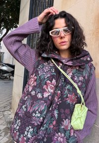 Γυναίκα με σγουρά σκούρα μαλλιά φορώντας λευκά γυαλιά ηλίου σε σχήμα γάτας, μοβ πλεκτό πουλόβερ, φλοράλ γιλέκο και κρατώντας μια μικρή πράσινη τσάντα, έξω.