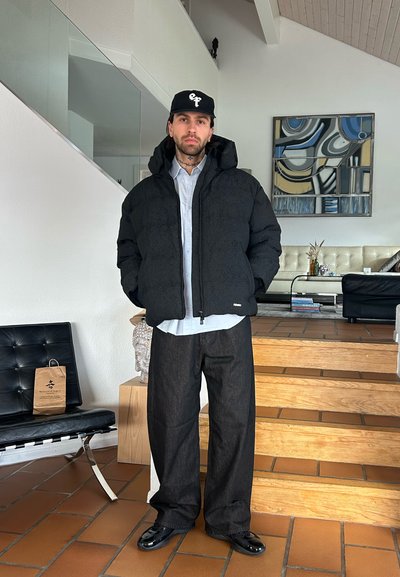 Hombre con chaqueta acolchada negra, gorra negra, pantalones anchos oscuros y zapatos negros de pie en el interior cerca de escaleras de madera y muebles modernos.