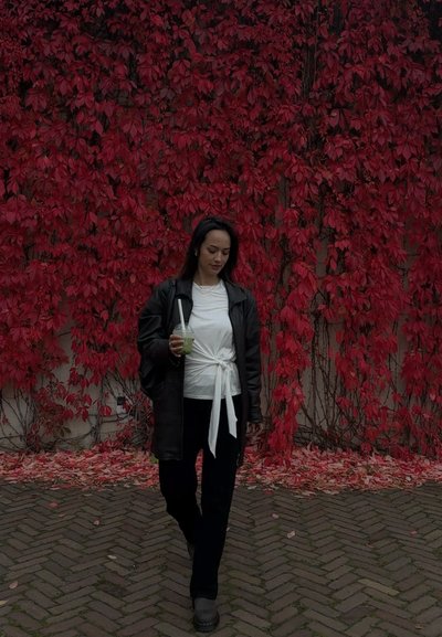 Chaqueta de cuero negra, top blanco anudado, pantalones negros. Sosteniendo una bebida verde, caminando contra un fondo de follaje rojo y un camino de ladrillos.