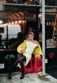 Kollane puffer-jakk valge triibulise kleidi, mustade sukkade ja pahkluu-saabastega. Istub punastel taburettidel, taga peegeldav klaaspind.