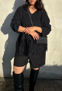 Frau trägt schwarzen Strickpullover mit Spitzenabschluss, schwarze Shorts, kniehohe Stiefel und kleine schwarze Umhängetasche, steht vor einer hellen Wand.