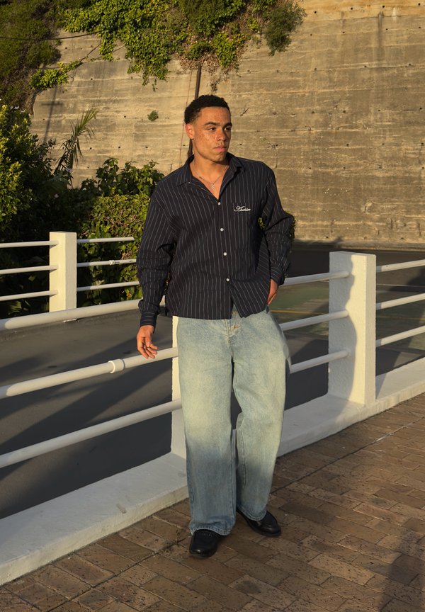 Homme debout près d'une rambarde blanche, portant une chemise rayée noire et un jean bleu clair ample, avec des plantes et un mur en béton en arrière-plan.