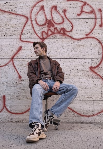 Chaqueta de cuero marrón, suéter de cuello alto gris, jeans azul claro y zapatillas beige. Sentado en una silla giratoria contra una pared de graffiti.