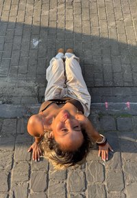 Een persoon in een bruine top en beige broek ligt op een gestructureerd grijs stenen oppervlak, terwijl zonlicht zachte schaduwen werpt.