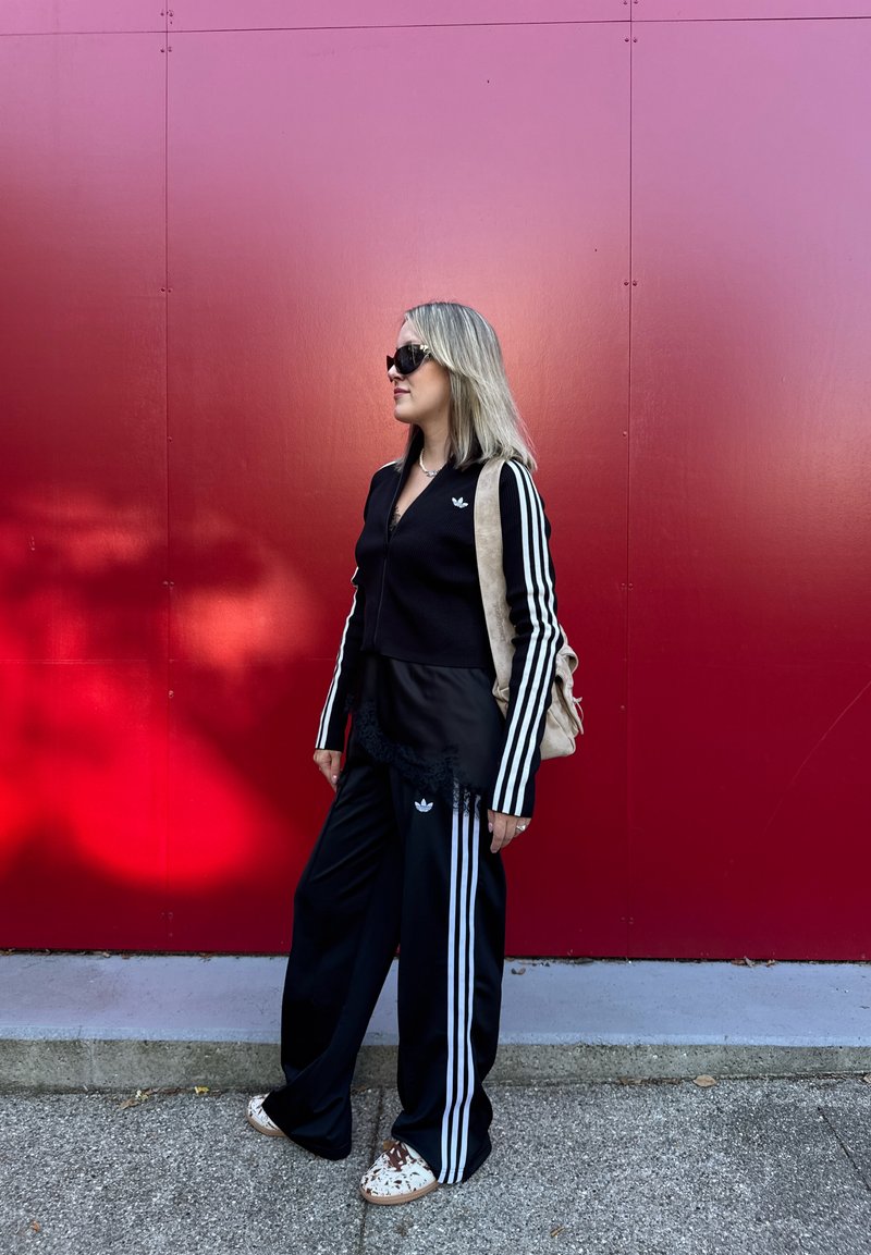Ensemble de sport noir avec des rayures blanches, comprenant une veste à zip côtelée et un pantalon à jambes larges. Sac à dos beige et chaussures à motif. Fond rouge.