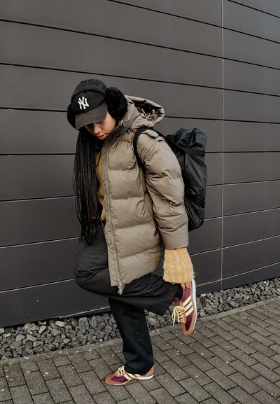 Chaqueta de plumas en gris claro, suéter de punto texturizado, gorra negra con orejeras, mochila negra, pantalones oscuros y zapatillas marrones/blancas.