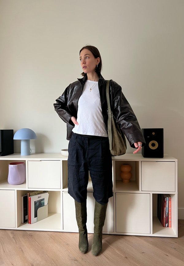 Donna con giacca di pelle nera, camicia bianca, pantaloni neri e stivali verdi, in piedi davanti a scaffali bianchi con libri, lampada e altoparlanti.