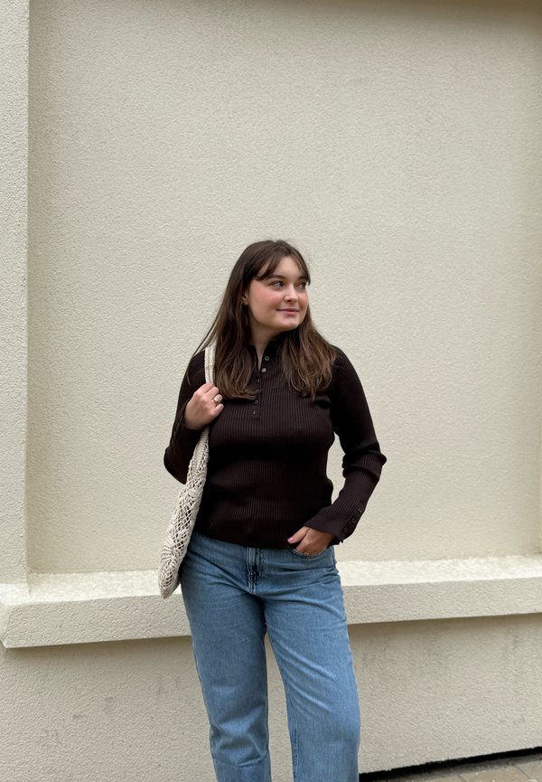 Jeune femme aux longs cheveux bruns se tient contre un mur beige, portant un haut noir à manches longues, un jean bleu, et porte un sac crocheté beige à l'épaule.