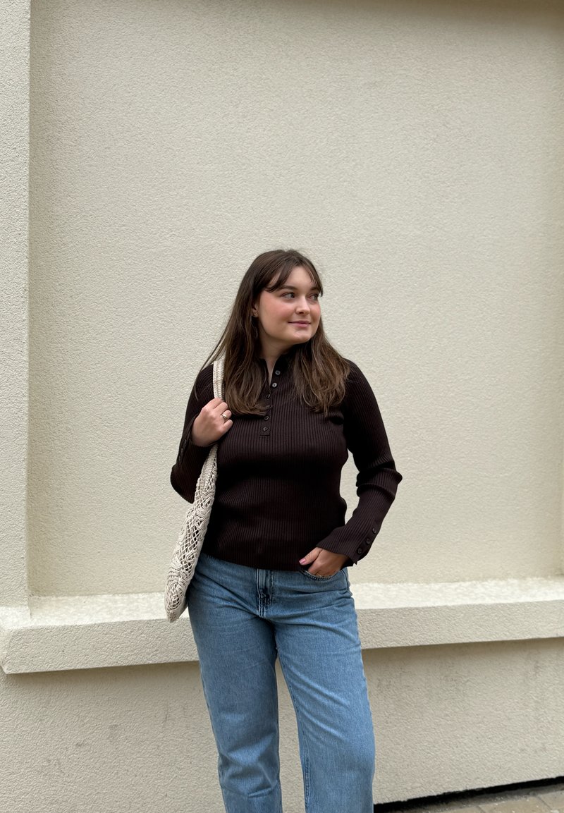 Jeune femme aux longs cheveux bruns se tient contre un mur beige, portant un haut noir à manches longues, un jean bleu et portant un sac en crochet beige à l'épaule.