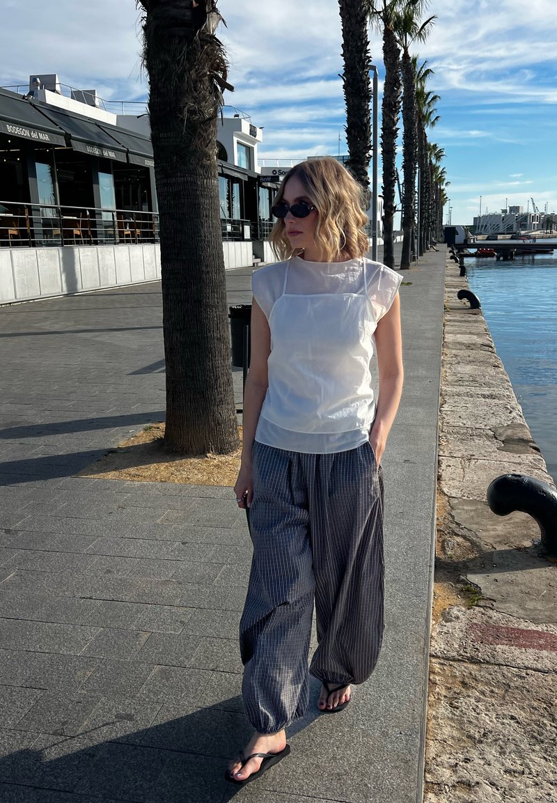 Frau mit Sonnenbrille, die ein weißes Oberteil und weite, gestreifte Hosen trägt, spaziert an einer von Palmen gesäumten Uferpromenade an einem sonnigen Tag.