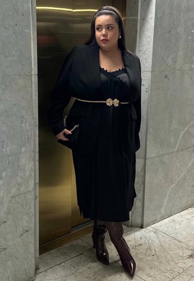 Blazer negro sobre un vestido negro, acentuado con un cinturón floral dorado. Botas de caña alta burdeos completan el look. Fondo de mármol.