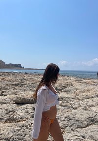 Biely, dlhý rukáv, otvorený top cez bikiny s farebným viazaním na boku; skalnatá pláž v popredí s pokojnou vodou a modrou oblohou na pozadí.