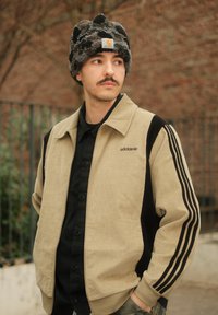 Veste en velours côtelé beige avec des manches noires, trois rayures noires sur chaque bras et un logo adidas sur le côté gauche. Chemise noire boutonnée en dessous.