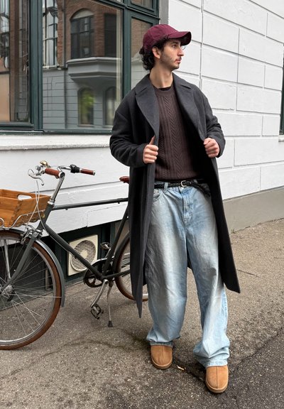 Joven con un abrigo largo y oscuro y una gorra burdeos de pie junto a una bicicleta con una cesta de madera en una acera de la ciudad.