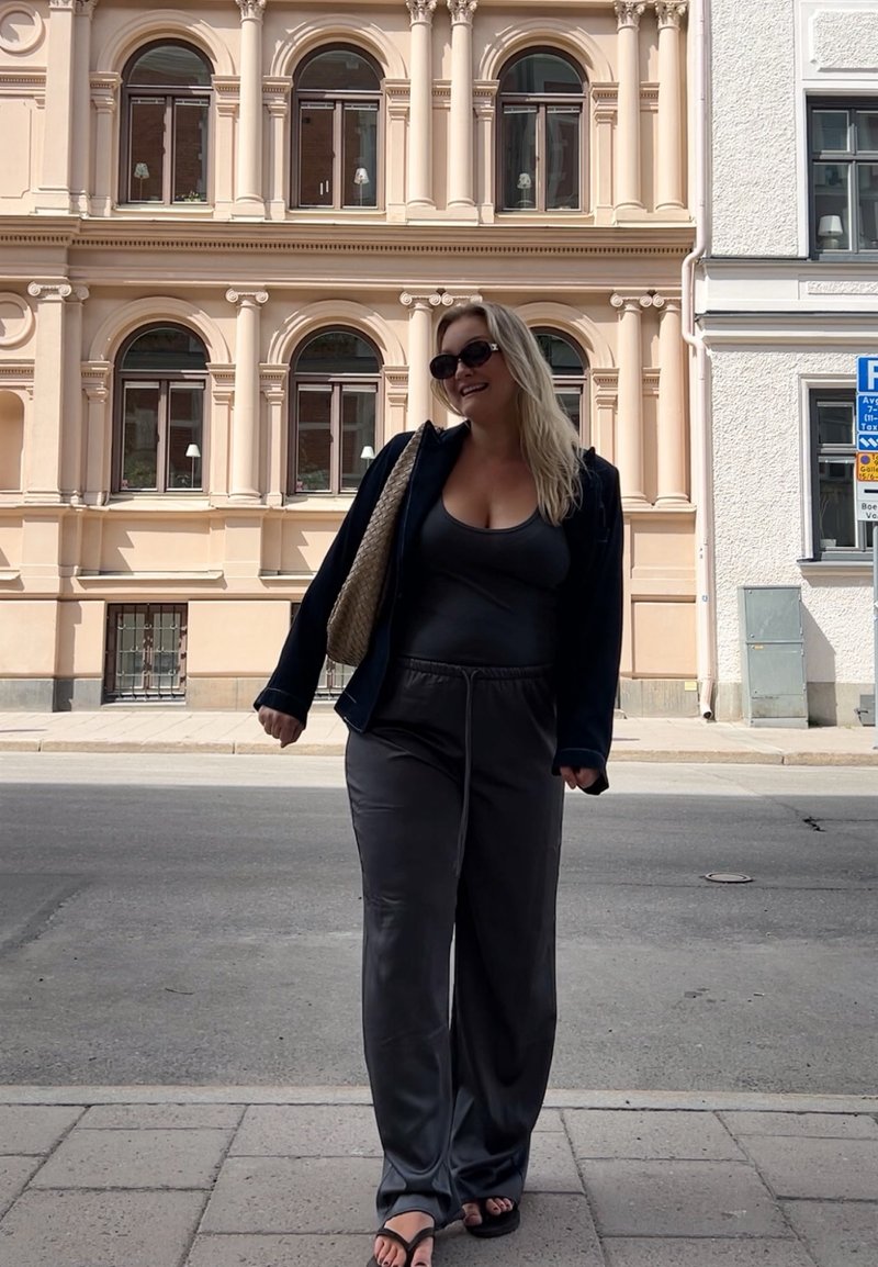 Ένα άτομο ντυμένο με ένα μαύρο σακάκι πάνω από ένα γκρι αμάνικο τοπ και γκρι φόρμα, κρατώντας μια υφαντή τσάντα και φορώντας μαύρες σαγιονάρες, στέκεται σε ένα πεζοδρόμιο.