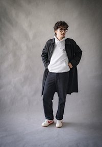 Czarny płaszcz na szarym hoodie z grafiką, ciemne dżinsy i białe sneakersy z czerwonymi akcentami. Szare tło o miękkiej teksturze.