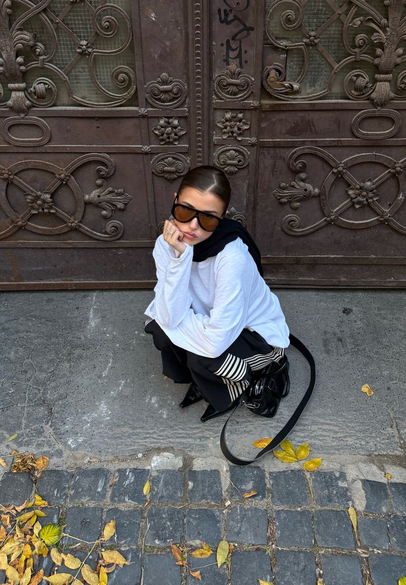 Kvinne med svarte solbriller og hvit topp som sitter på huk på brosteinsgate med høstløv, dekorativ metallport i bakgrunnen.