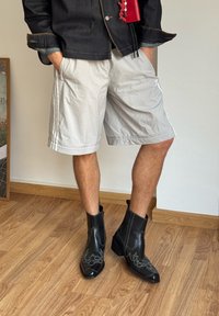 Czarne skórzane botki za kostkę z ostrymi noskami i dekoracyjnym szarym przeszyciem, noszone z jasnoszarymi szortami, ustawione na drewnianej podłodze.