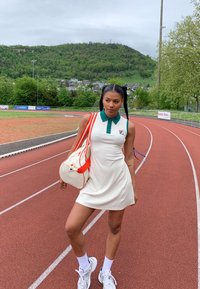 Robe polo blanche à côtes avec un col vert, arborant un logo Fila. Sac crème avec des sangles rouges et des baskets blanches. Piste d'athlétisme en arrière-plan.