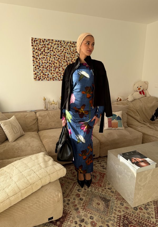 Femme portant un foulard beige, une robe bleue fleurie, une veste noire et des talons, tenant un sac à main noir dans un salon moderne.