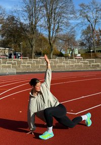 Idrottare i en ljusgrå jacka och svarta leggings utför en stretch på en röd löparbana. Ljusa sneakers med gröna detaljer och vita strumpor.