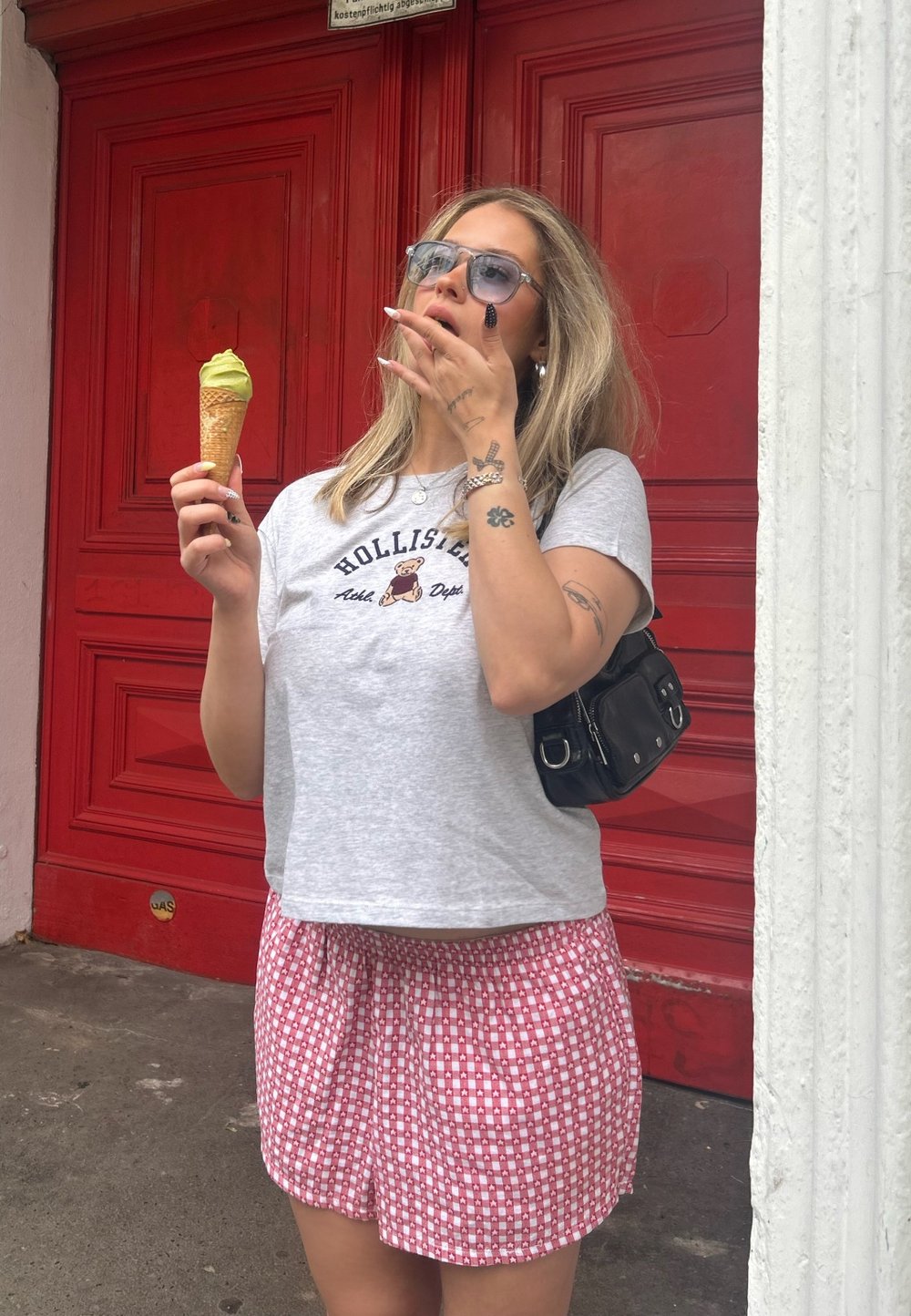 Grå t-shirt med ett björnmotiv, röd rutig shorts, håller en grön glasskonus, framför en klar röd dörr. Svart axelväska.