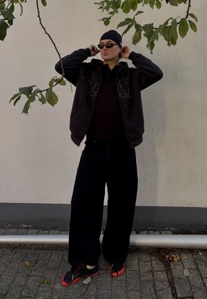Svart zip-up hoodie med stort logotyp, svart polo skjorta, breda svarta byxor och svarta/röda femtåiga skor. Bakgrunden har en enkel vägg och grönska.