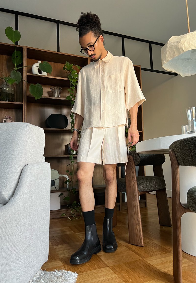 Homme aux cheveux bouclés et lunettes, portant une chemise beige ample et un short avec des chaussettes noires et des bottes, debout dans le salon près d'une table en bois et des chaises.