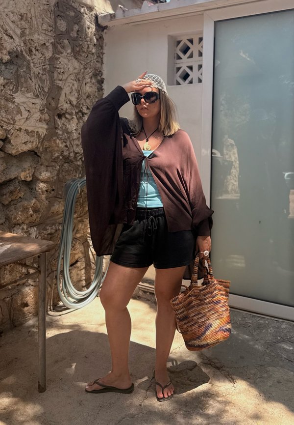 Femme portant des lunettes de soleil, un châle marron, un short noir et des tongs, tenant un sac tissé, debout dehors près d'un mur en pierre et d'une porte en verre.