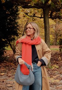 Beige trenchcoat, oransje skjerf, marineblå genser, lyseblå jeans, teksturert grå håndvevd veske med flettet design, høstblader i bakgrunnen.