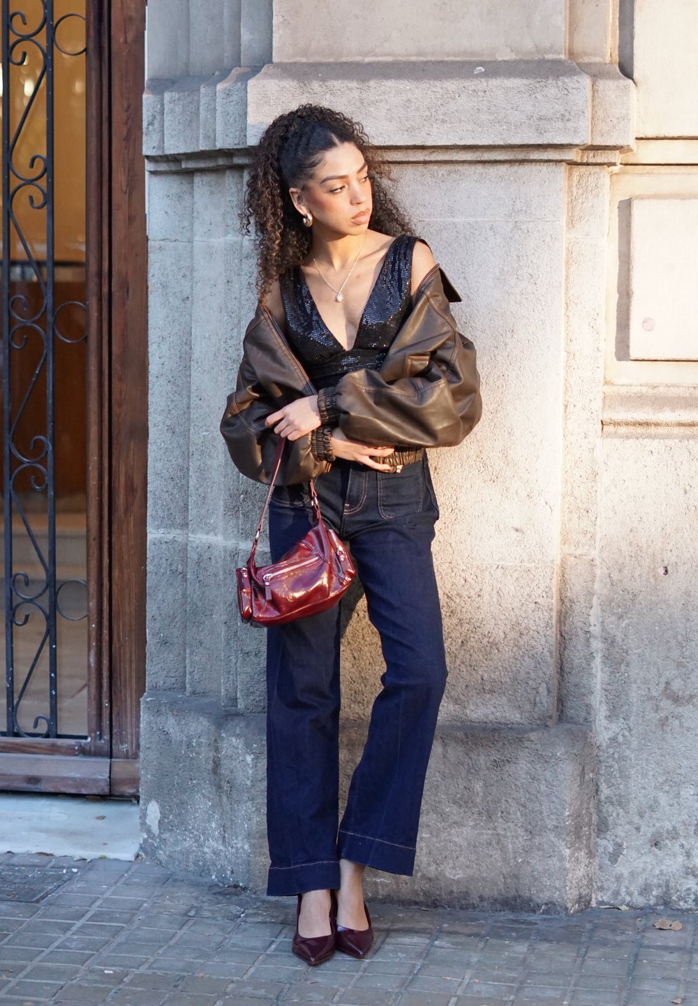 Braune übergroße Lederjacke, schwarzes Pailletten-Top, dunkle Jeans, burgunderroter Handtasche und burgunderrote High Heels, posiert vor einer Steinmauer.