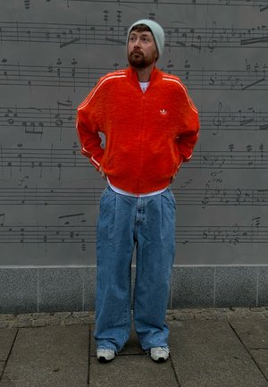 Chaqueta naranja con cremallera de textura peluda y rayas blancas, pantalones sueltos de mezclilla azul, camiseta blanca y gorro gris; de pie frente a un fondo de notas musicales.