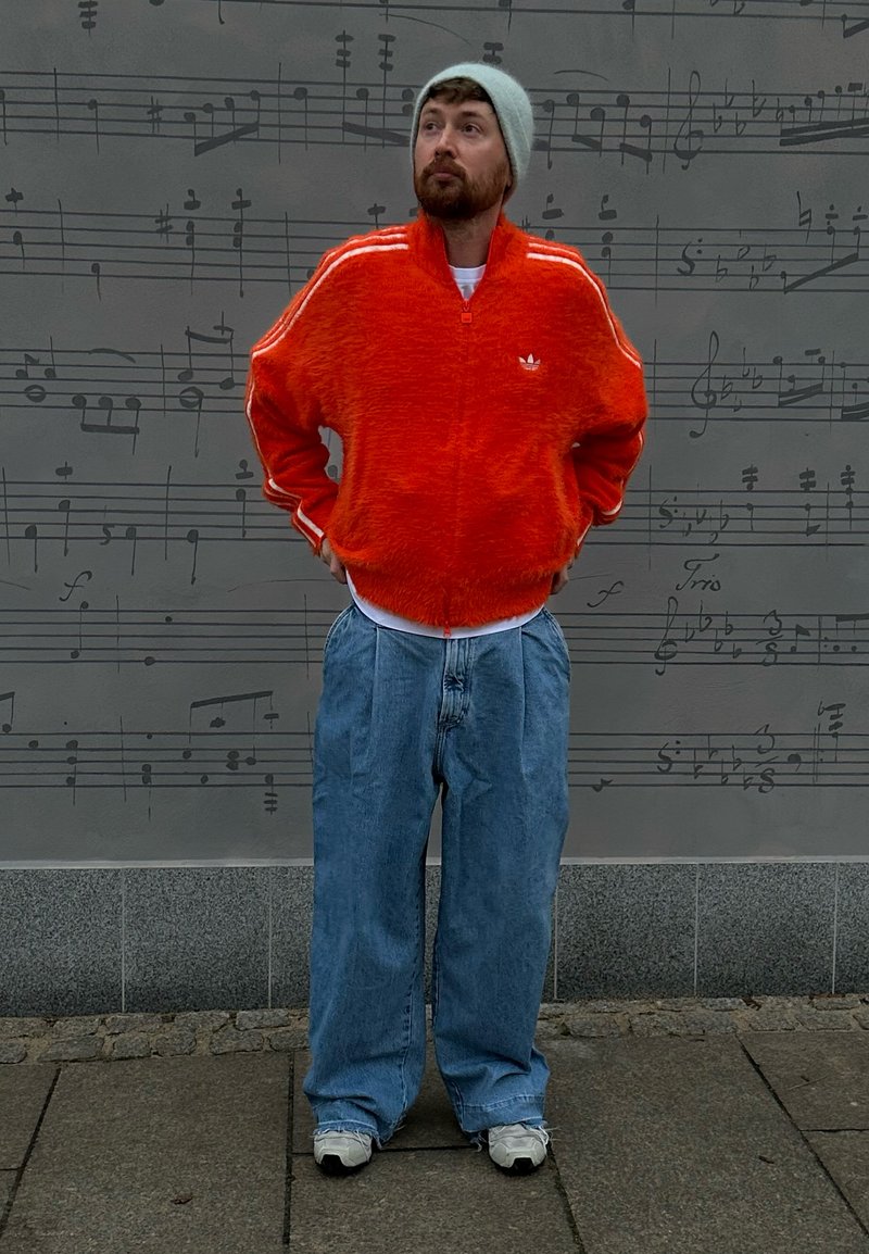 Orangene, flauschige Jacke mit Reißverschluss und weißen Streifen, lockere blaue Jeanshose, weißes T-Shirt und graue Mütze; vor einem Hintergrund mit Musiknoten stehend.