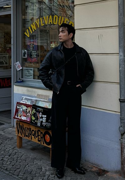 Chaqueta de cuero negra, suéter de cuello alto negro y pantalones de pierna ancha, llevados por una persona que está de pie junto a una tienda de discos con portadas de álbumes coloridas expuestas.