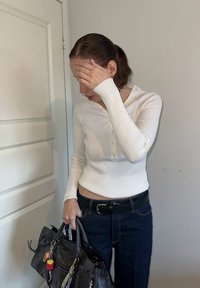 Bela rebrasta dolga majica s kratkimi rokavi henley z gumbi na vratu, v kombinaciji s temnimi kavbojkami in sivo torbico, ki ima barvite obeske.