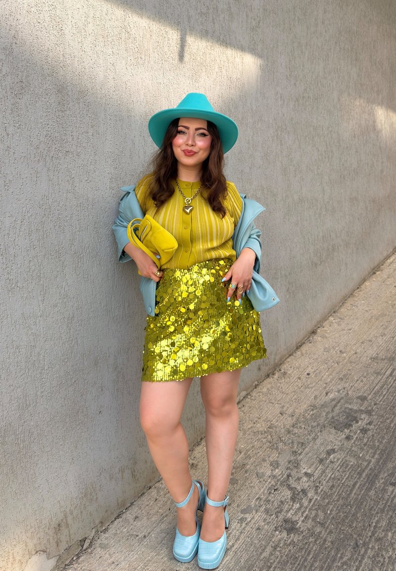Woman wearing a turquoise hat and jacket, yellow striped blouse, gold sequin skirt, and turquoise heels, standing against a concrete wall.