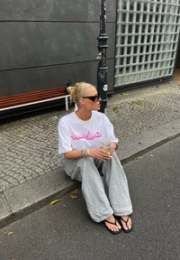 Άσπρη oversize μπλούζα με ροζ κείμενο, γκρι παντελόνι φόρμας και μαύρες παντόφλες. Κρατάει ένα διάφανο ποτήρι, καθισμένος σε μια λιθόστρωτη οδό.