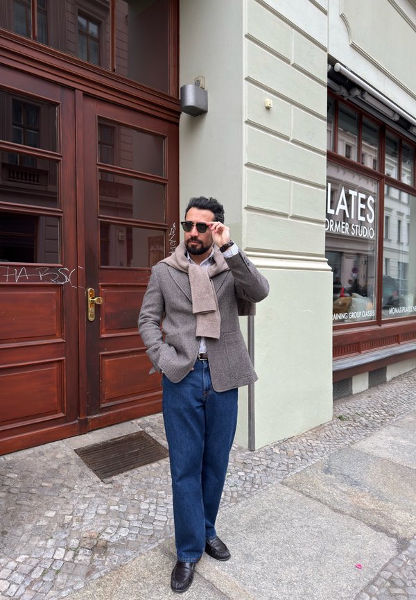 Muškarac sa sunčanim naočalama, sivim sakoom, bež šal i plavim trapericama stoji na kaldrmisanoj ulici blizu izloga pilates studija.