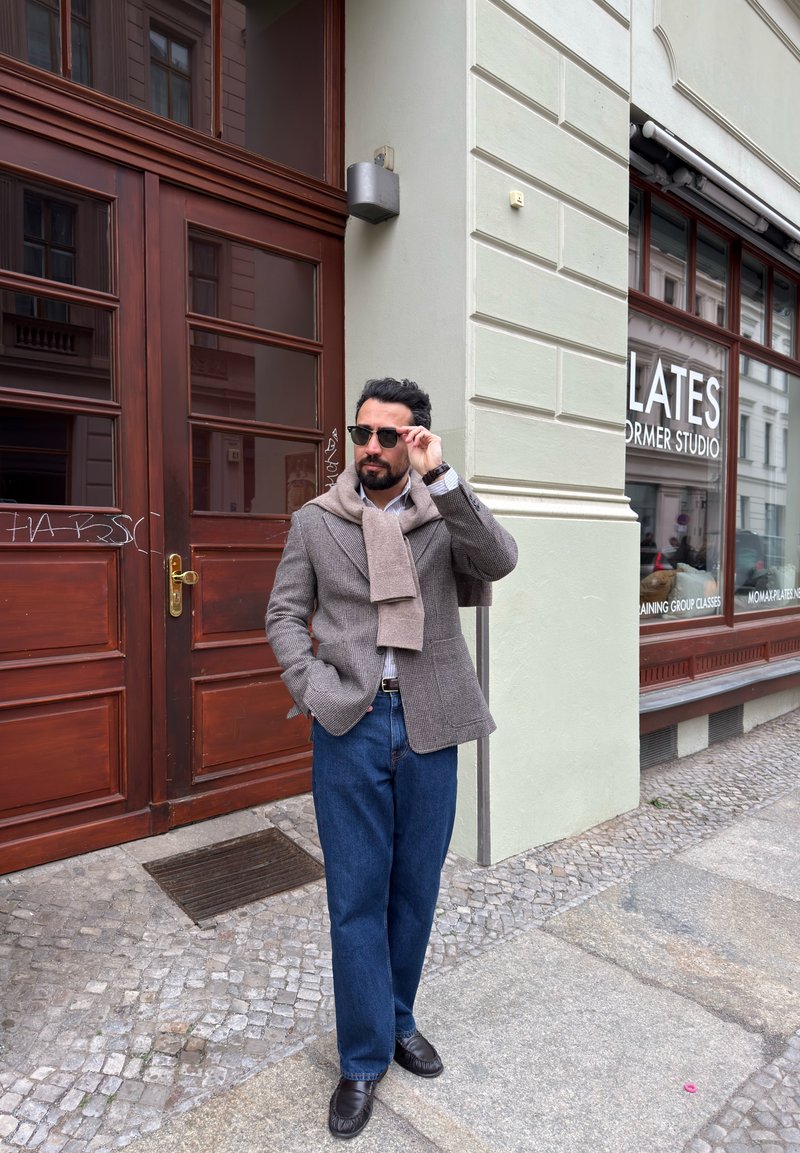 Homme portant des lunettes de soleil, un blazer gris, une écharpe beige et un jean bleu, debout dans une rue pavée près d'une vitrine de studio de Pilates.