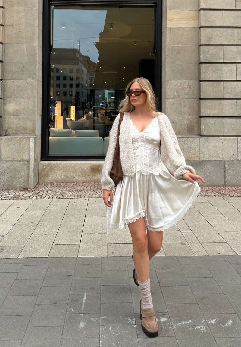 Abito bianco con pizzo, cardigan beige lavorato a maglia, borsa marrone e scarpe platform taupe abbinate a calzini chiari. Sfondo urbano.