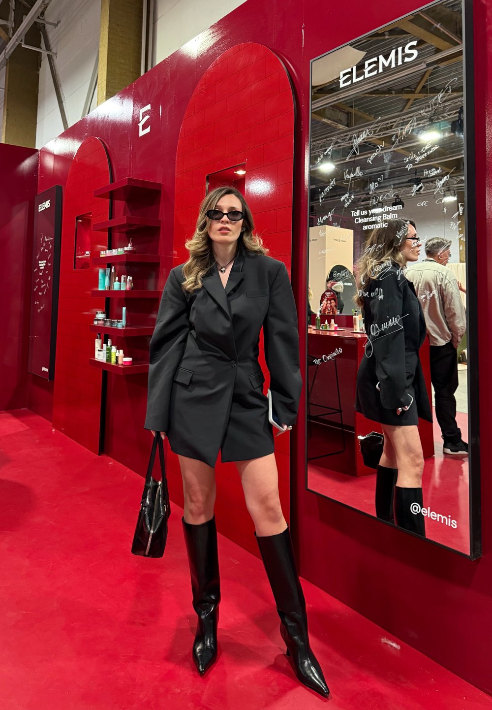Blazer noir oversize, bottes noires à talons hauts, lunettes de soleil élégantes. Fond en carreaux rouges avec présentoirs de produits et miroirs.