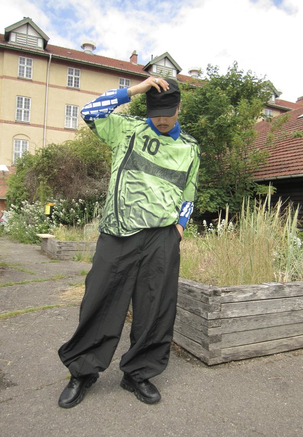 Osoba koja nosi zelenu i plavu sportsku majicu, crne široke hlače i crnu kapu na otvorenom blizu drvenih žardinjera i zgrada.