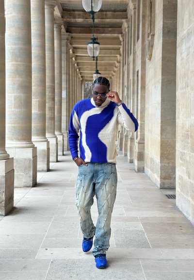 Jersey de punto en azul y crema con diseño abstracto; vaqueros holgados azul claro con detalles texturizados; zapatillas azules; gafas de sol puestas.